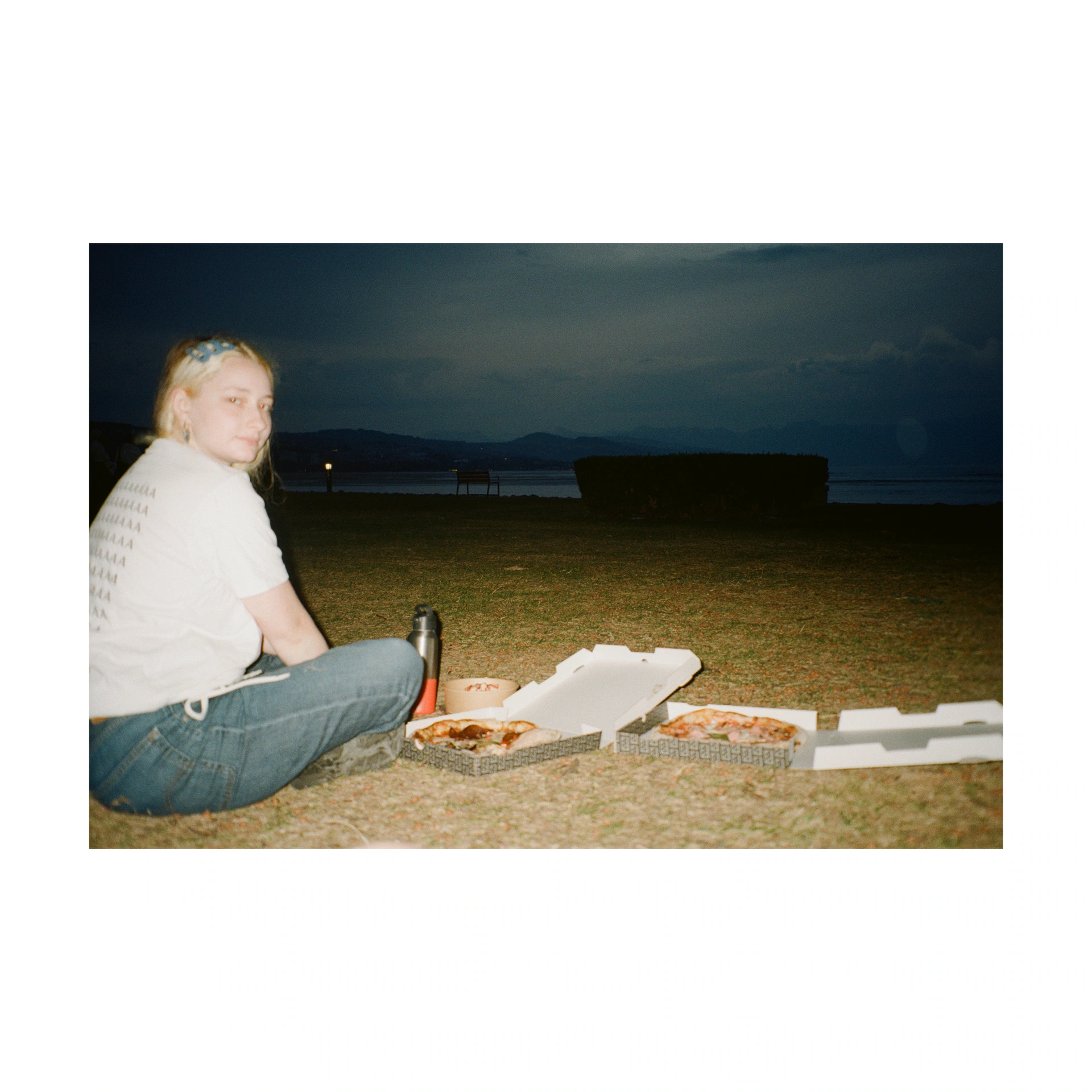 Person sitting on grass beside a pizza box at dusk